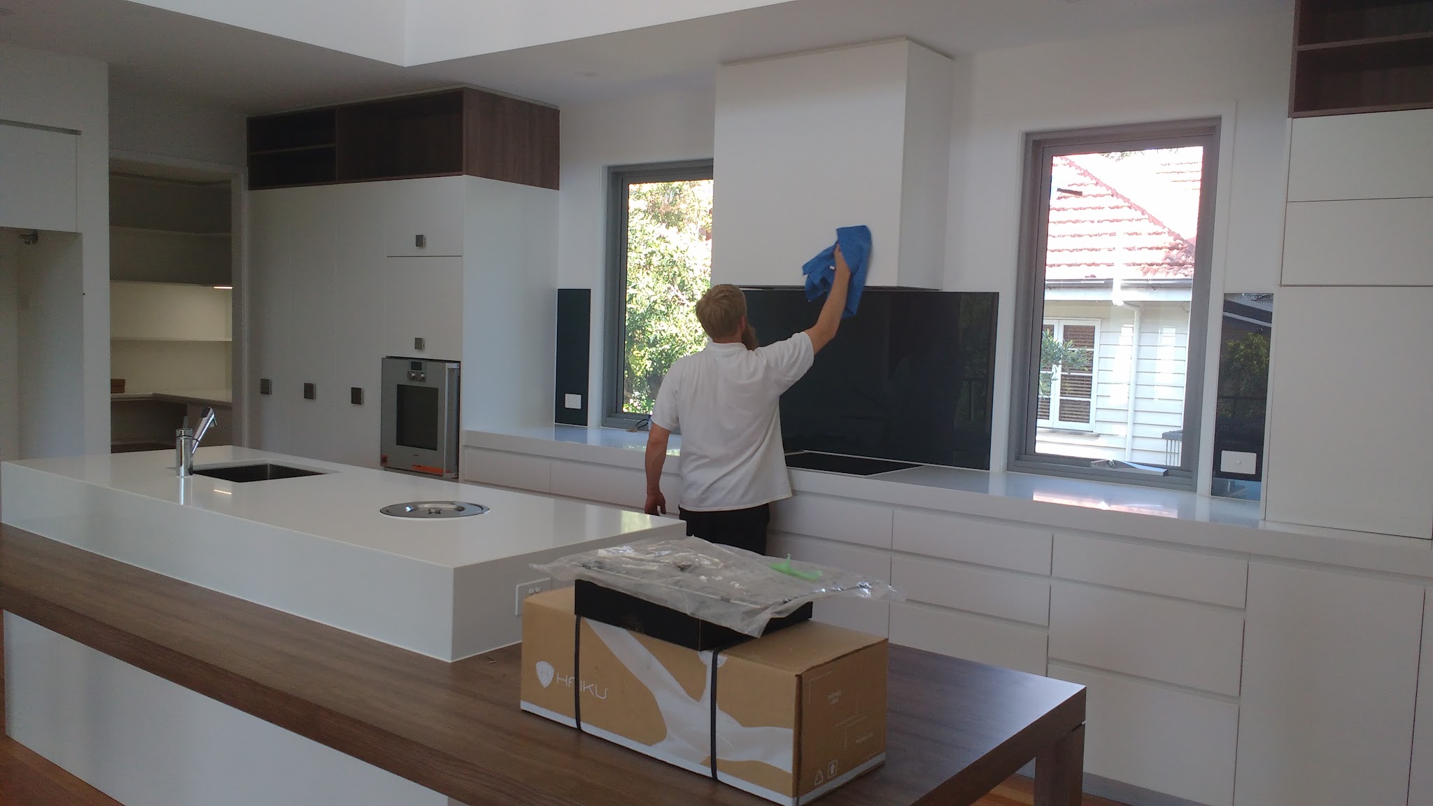 Cleaner polishing the rangehood above a black glass splashback in a large modern kitchen